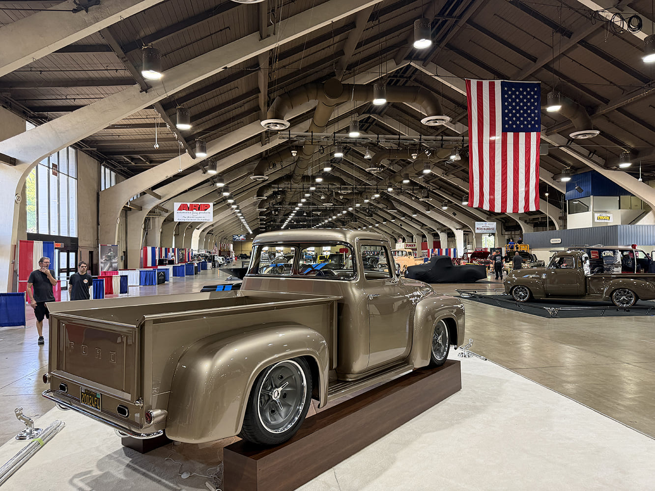 High rear three-quarter view of a champagne-bronze 1956 Ford F-100 custom pickup displayed on a wooden riser beneath the American flag inside Fairplex Expo Hall.