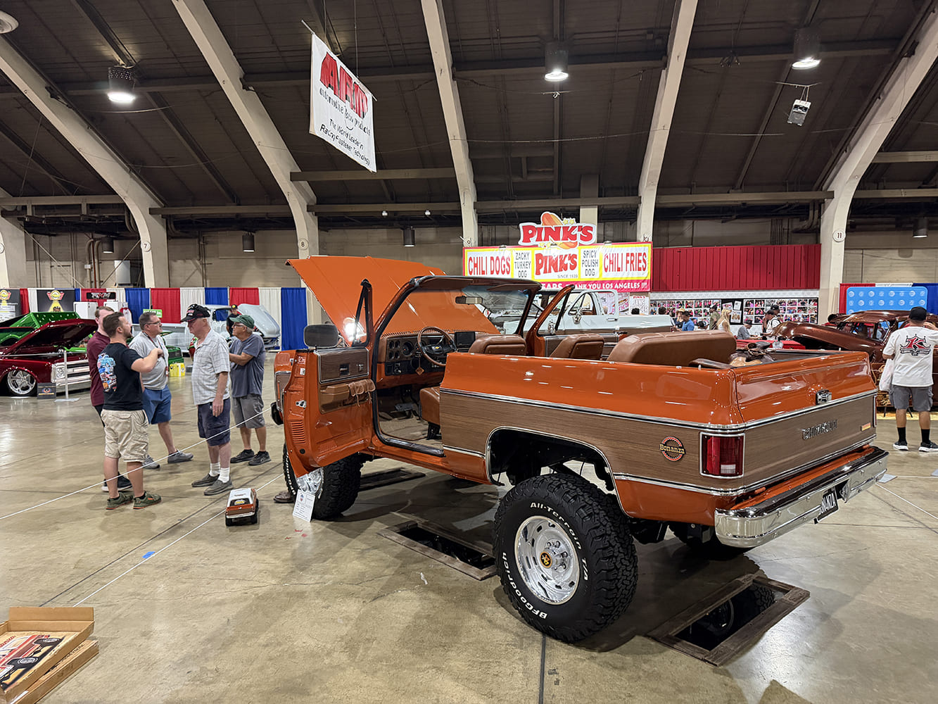 Rear three-quarter perspective of a lifted copper-orange Chevrolet K5 Blazer with woodgrain paneling, open doors, and all-terrain tires displayed near the Pink's Chili Dogs booth.
