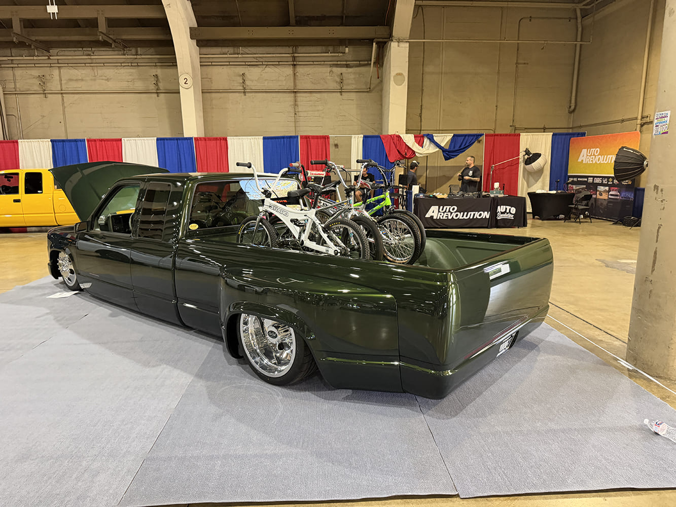 Low-angle rear three-quarter view of an ultra-low dark metallic green Chevrolet C3500 dually pickup carrying vintage BMX bicycles in the bed at the Auto Revolution booth.
