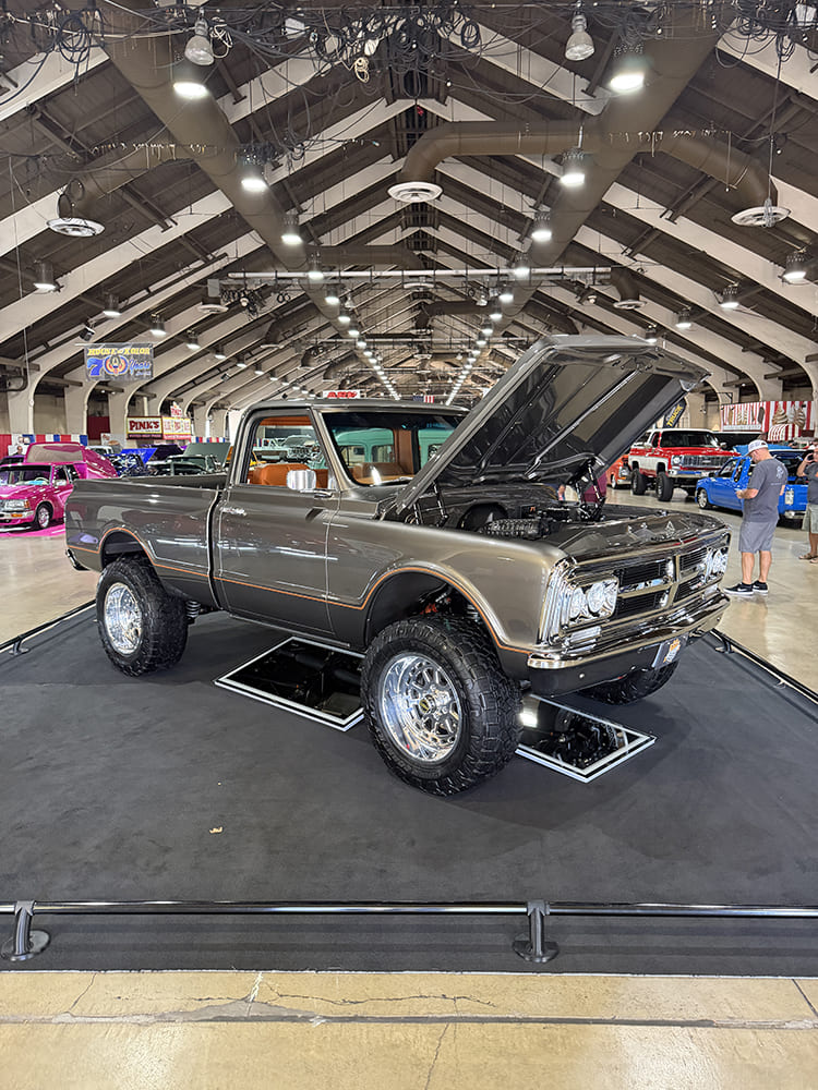 Front three-quarter angle of a metallic pewter-gray lifted 1967 Chevrolet 4x4 pickup truck with an open hood and polished off-road wheels displayed over undercarriage mirrors.
