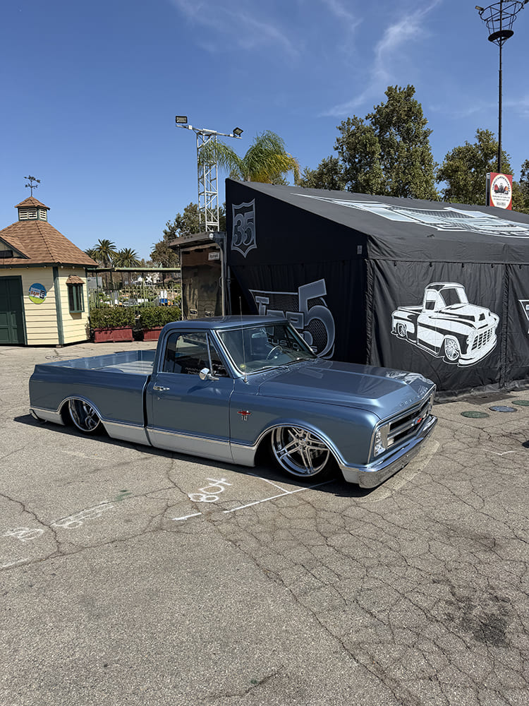 Low front three-quarter view of a lowered slate blue Chevrolet C10 pickup laid out flat on the asphalt outside a black event canopy at the Fairplex grounds.