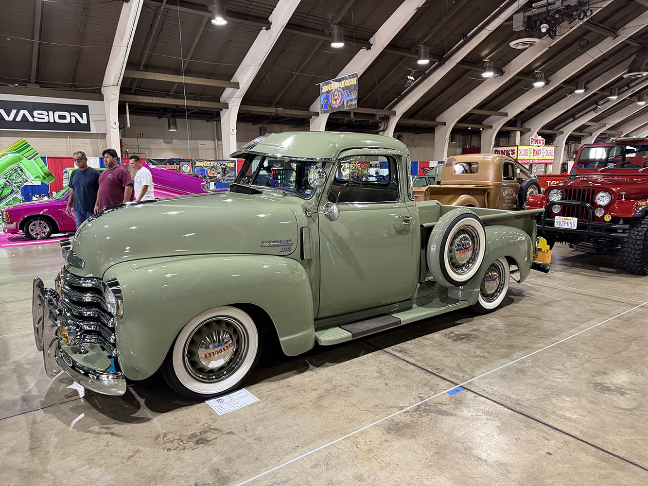 Front three-quarter perspective of an original-style pastel sage green Chevrolet 3100 pickup truck with cab sun visor, side-mount spare, and wide whitewall tires on the showroom floor.
