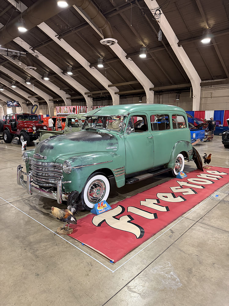 Front three-quarter view of a weathered patina green Chevrolet Suburban Carryall parked on a red Firestone display rug with decorative roosters inside Expo Hall.