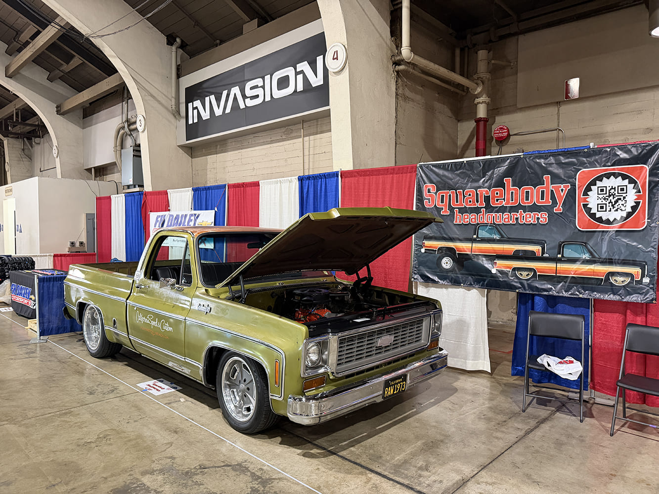 Front three-quarter angle of an olive green Chevrolet squarebody pickup truck with an open hood on display in front of the Squarebody Headquarters vendor banner.