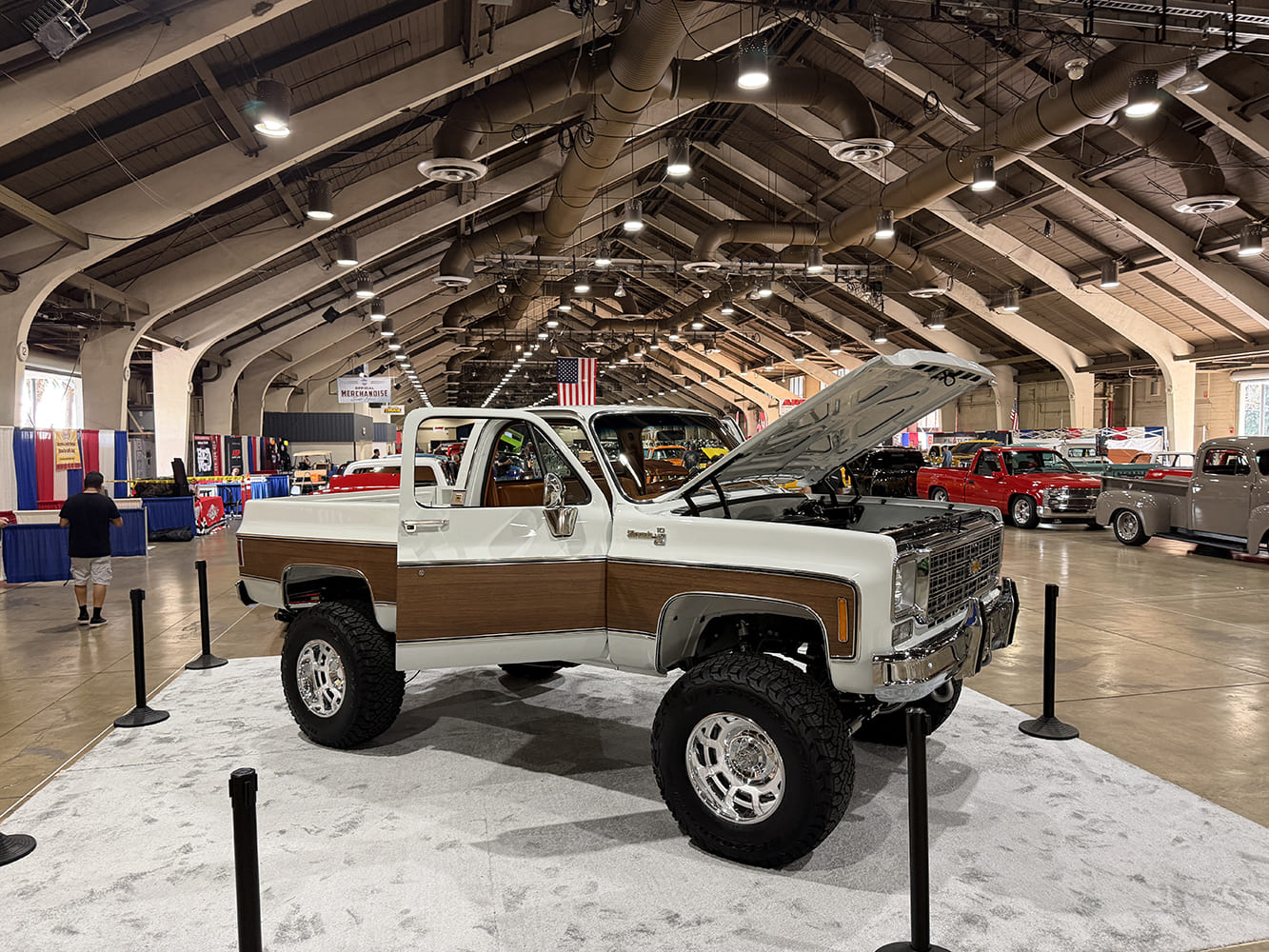 Front three-quarter view of a pristine white lifted squarebody Chevrolet K10 Silverado pickup featuring woodgrain exterior trim, tan interior, and massive off-road tires on white carpet.