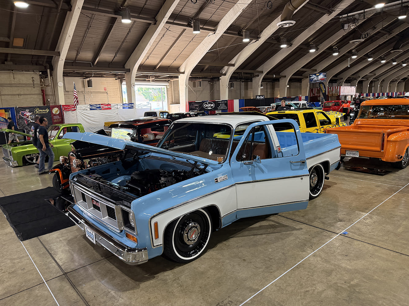 Front three-quarter angle of a lowered light blue and white two-tone squarebody GMC pickup with an open hood and driver door inside Expo Hall at the Grand National Truck Show.