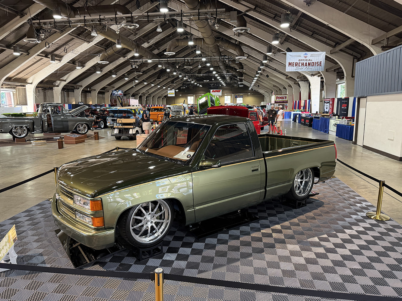Elevated front three-quarter view of a lowered dark olive metallic Chevrolet C1500 OBS pickup with polished multi-spoke wheels on a checkered floor display.