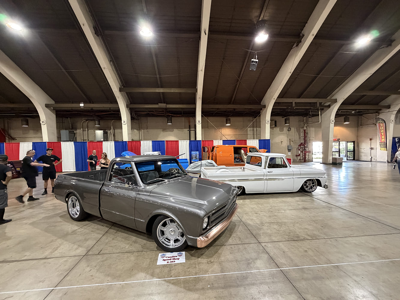 Front three-quarter perspective of a metallic pewter-gray Chevrolet C10 pickup with copper front bumper accents parked alongside a lowered white C10 in an expo hall.