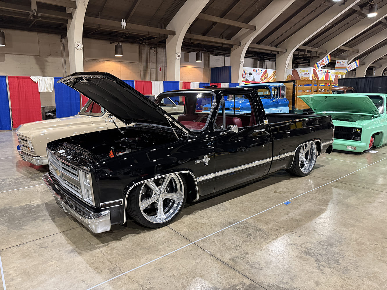 Front three-quarter view of a lowered jet-black Chevrolet Silverado squarebody pickup with an open hood showing a detailed engine bay in an indoor car show row.