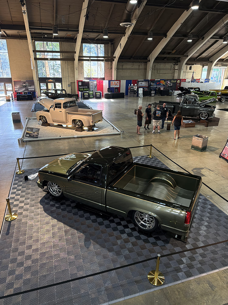 Elevated looking down at an olive-metallic custom pickup displaying custom wheel tubs, smooth bed floor, and chrome-accented frame inside the convention center.