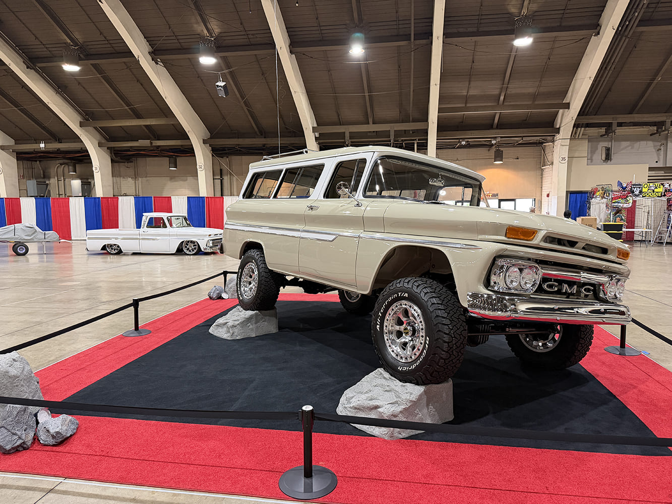 Low-angle front three-quarter view of a lifted tan vintage GMC Suburban 4x4 posed with wheels articulated over artificial boulders on red carpet.