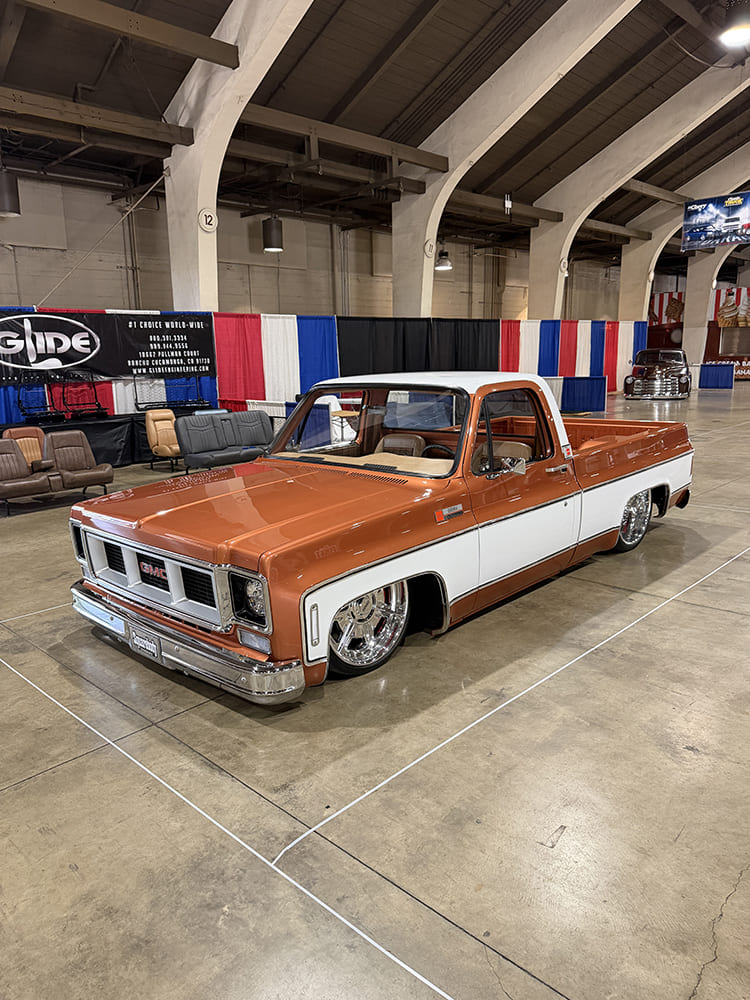 High front three-quarter perspective of a lowered two-tone metallic copper and white squarebody GMC pickup sitting low over polished chrome wheels.