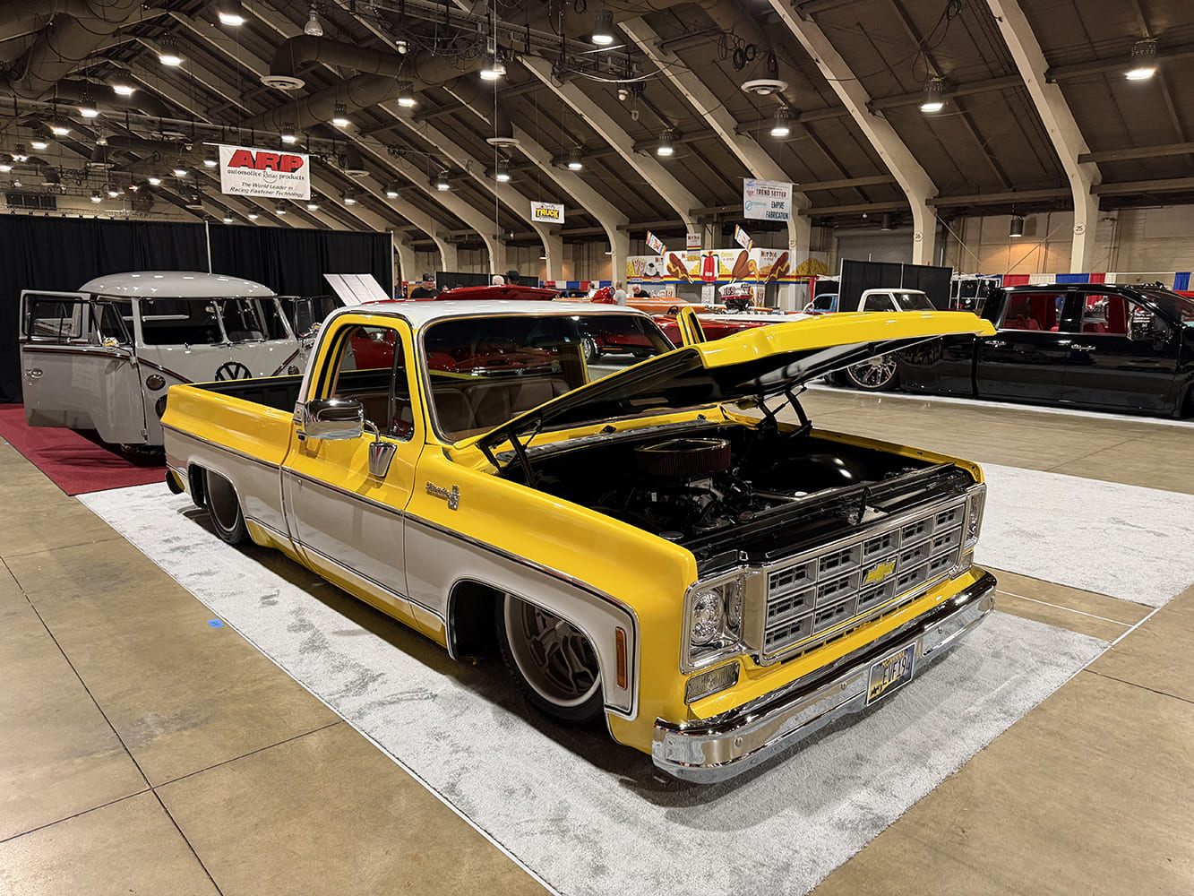 Front three-quarter view of a slammed yellow and white Chevrolet C10 pickup with an open hood displaying a performance V8 on a white carpet pad.