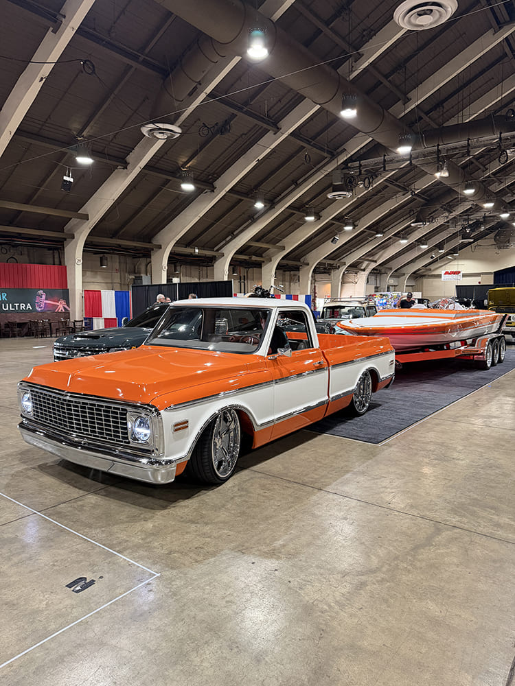 Front three-quarter view of a lowered orange and white Chevrolet C10 pickup hitched to a color-matched custom speedboat and trailer inside the exhibition building.