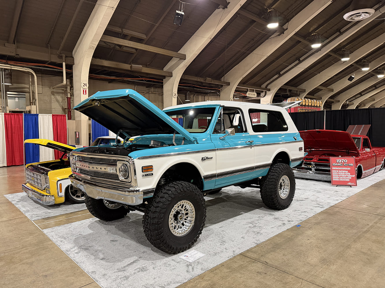 Front three-quarter angle of a lifted turquoise and white two-tone 1970 Chevrolet K5 Blazer with open hood on polished beadlock off-road wheels at the Grand National Truck Show.