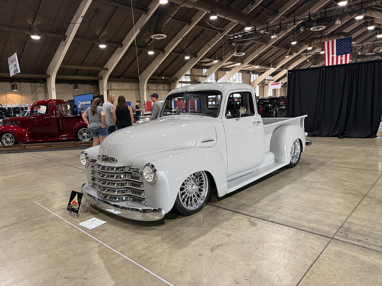 Front three-quarter view of a lowered glossy light gray Chevrolet 3100 pickup truck resting on intricate chrome wire wheels inside the expo hall.