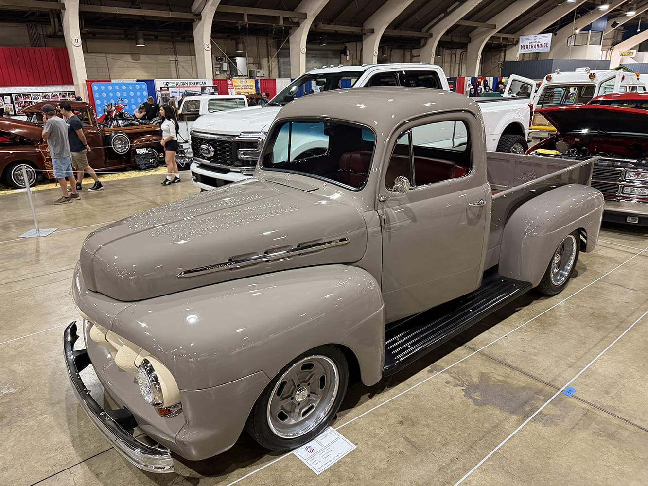 High front three-quarter view of a glossy warm gray 1949 Ford F-1 pickup with a louvered hood and bronze-finish wheels on the show floor.
