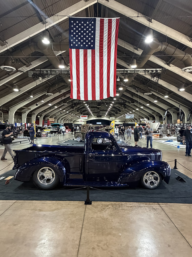 Driver-side profile of a lowered dark midnight blue 1940 Ford pickup truck parked on display carpet directly beneath a large hanging American flag.