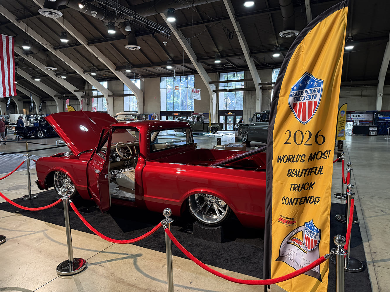 Rear three-quarter angle of a slammed metallic crimson 1972 Chevrolet C10 pickup with open hood and door behind a yellow World's Most Beautiful Truck Contender banner.