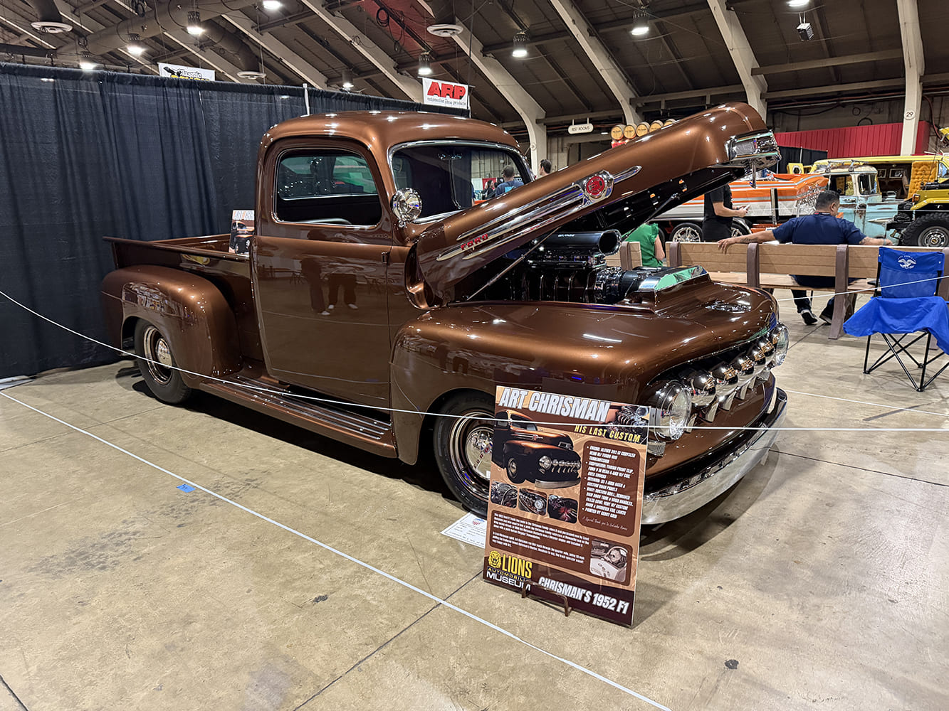 Front three-quarter view of a metallic root beer brown 1952 Ford F-1 pickup with tilt front hood displaying the build history board from the Lions Automobilia Museum.