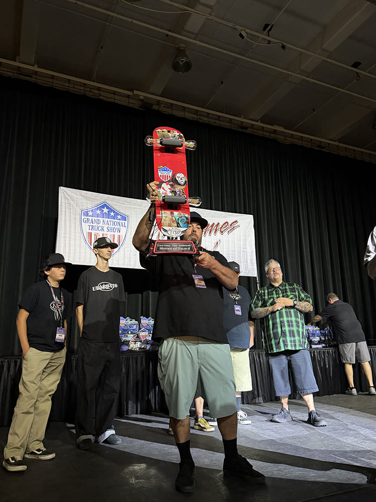 Award recipient holding up a custom red painted skateboard deck trophy on stage alongside fellow participants at the Grand National Truck Show awards ceremony.