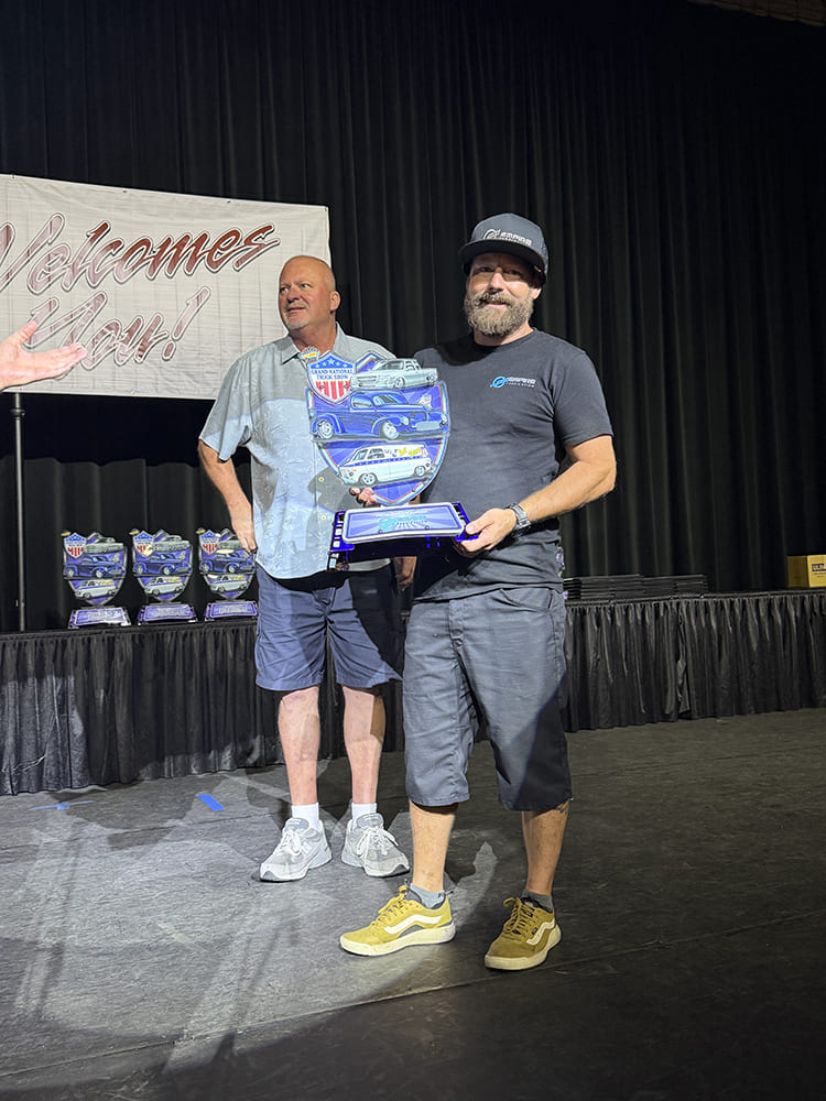 An award recipient wearing a cap and beard proudly holding a blue acrylic truck trophy beside the event presenter on the Grand National stage.