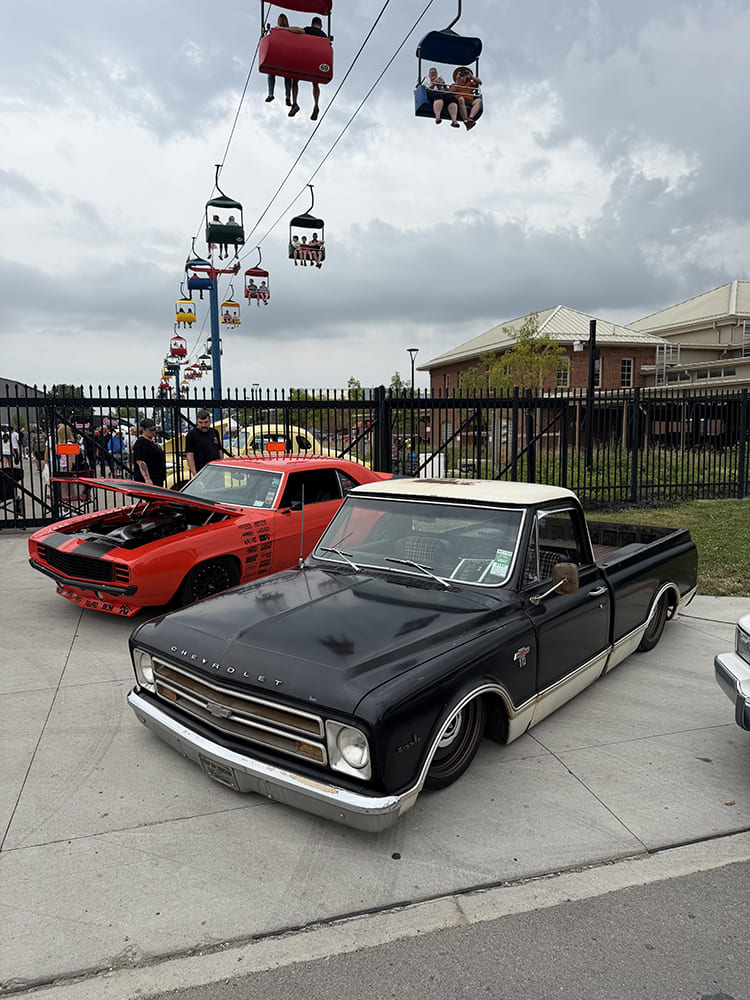 Front three-quarter view of a lowered black and white two-tone 1967 Chevrolet C10 pickup parked beside an orange Camaro beneath skyride cable cars at Goodguys Columbus.