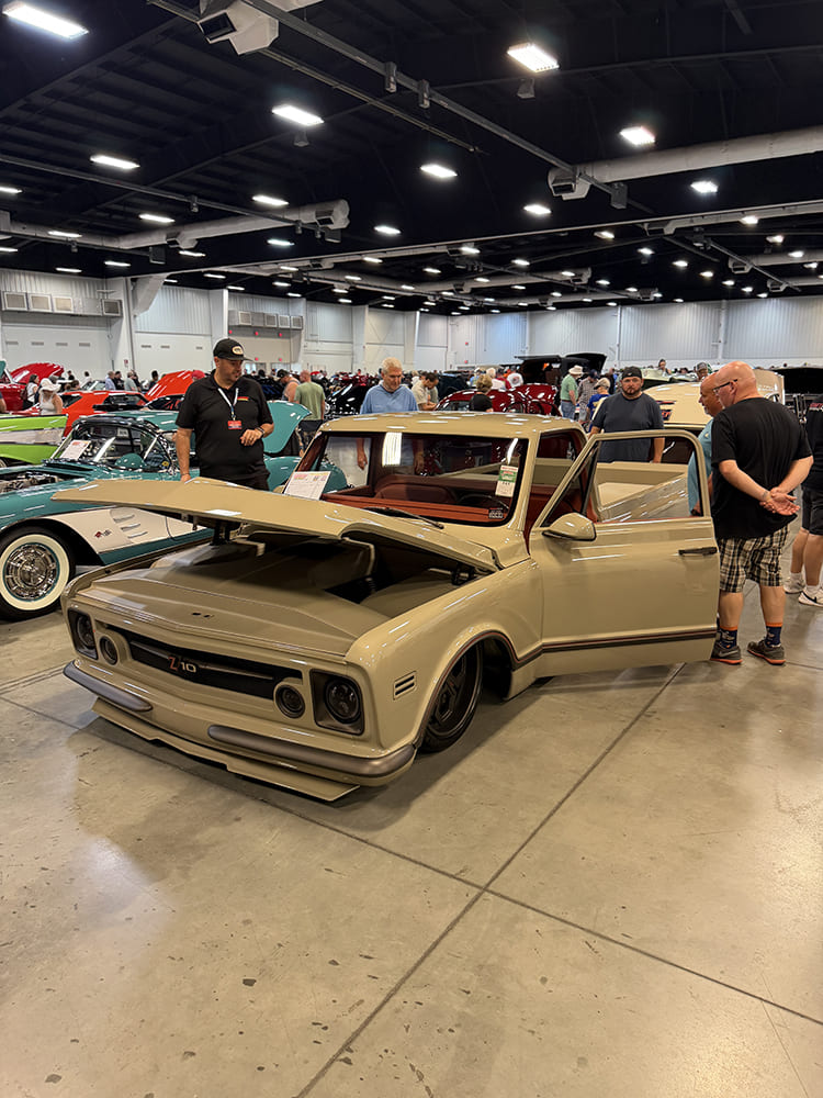 High front three-quarter view of a fully shaved tan custom Chevrolet C10 pickup with open hood, flush bumper, and brown interior displayed inside the convention hall.