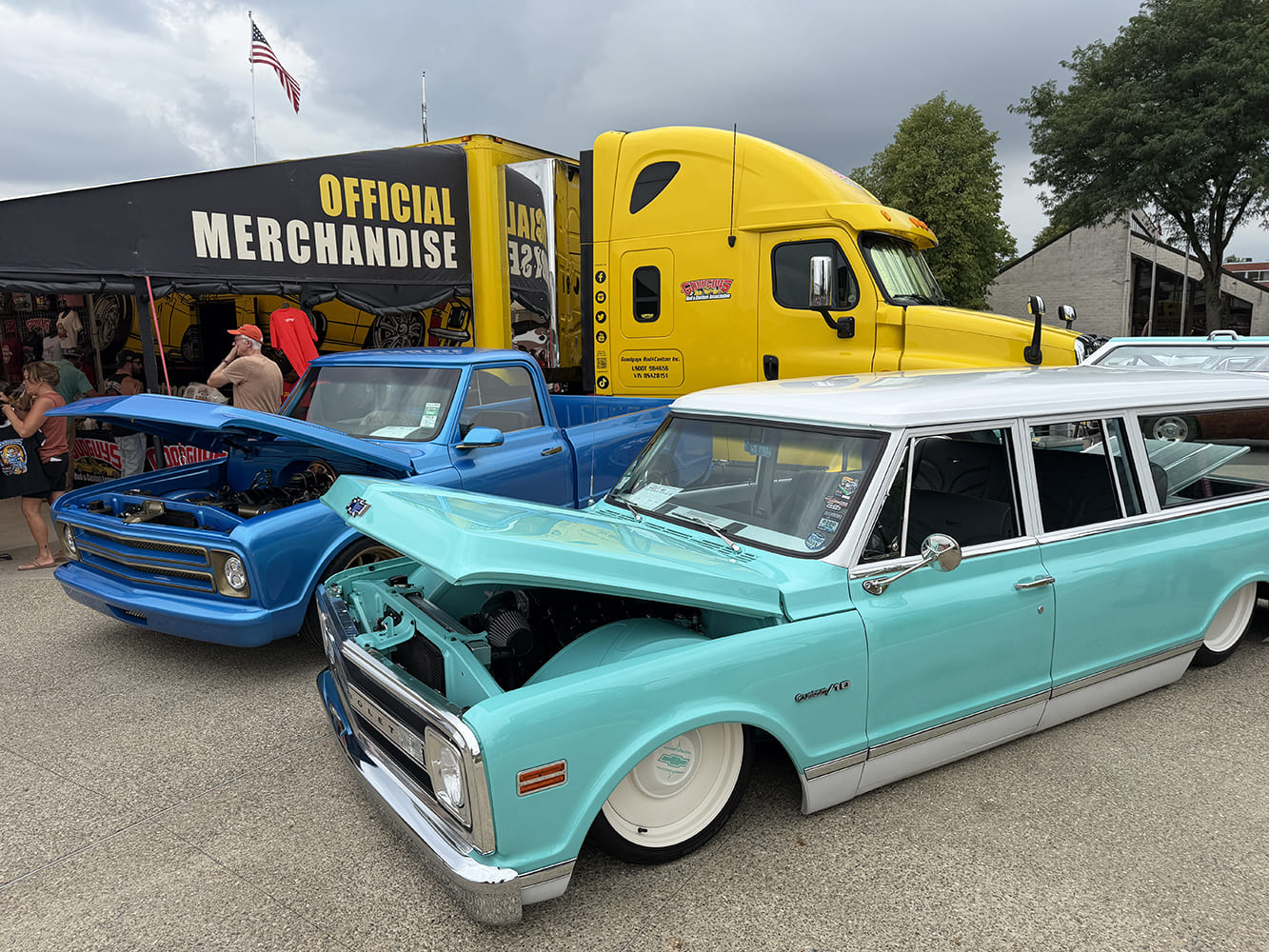 Front three-quarter angle of a slammed two-tone seafoam turquoise and white Chevrolet Suburban parked next to a bright blue C10 pickup outside the yellow Goodguys merchandise rig.