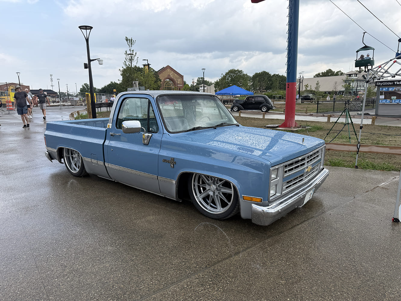 Front three-quarter perspective of a lowered light blue and silver two-tone Chevrolet Silverado squarebody pickup parked on wet asphalt beneath the fairgrounds skyride.