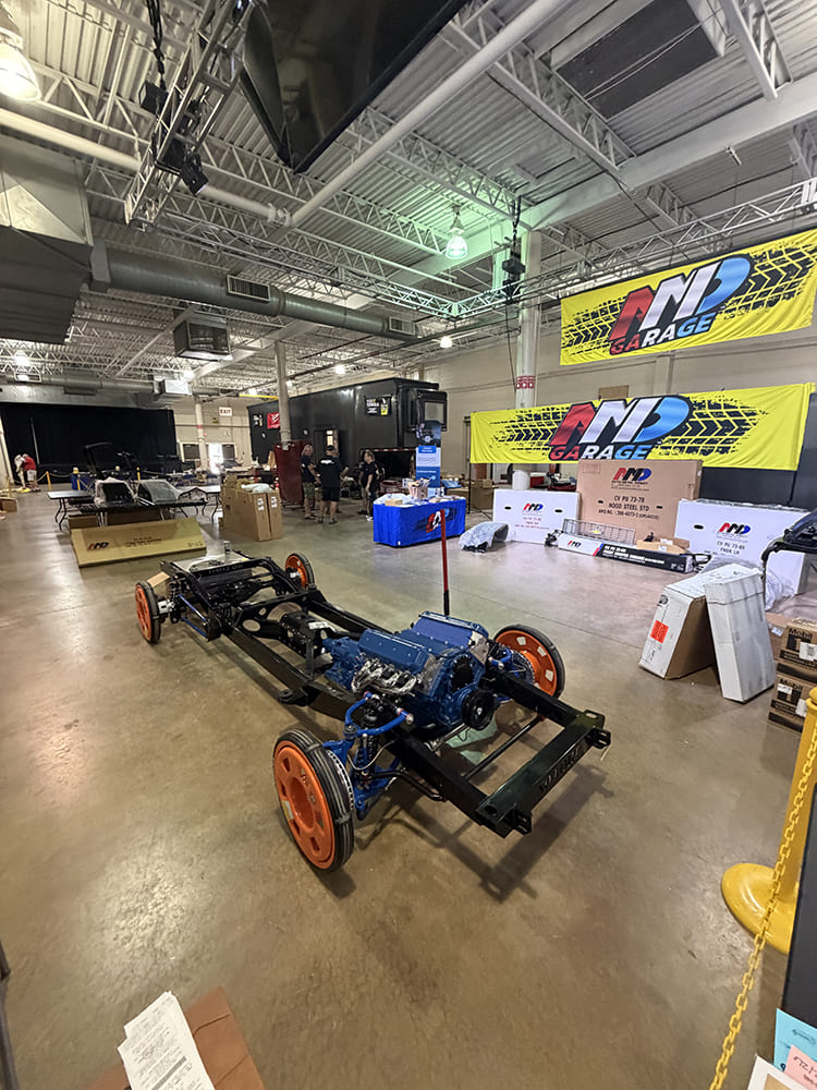 High-angle overview of a custom truck chassis equipped with a blue V8 engine, orange transport wheels, and performance coilover suspension inside an exhibition hall.