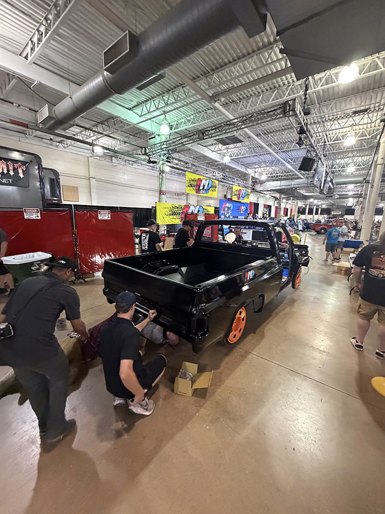 Rear three-quarter perspective of builders mounting a black steel fleetside bed onto a custom truck chassis inside an indoor expo hall.