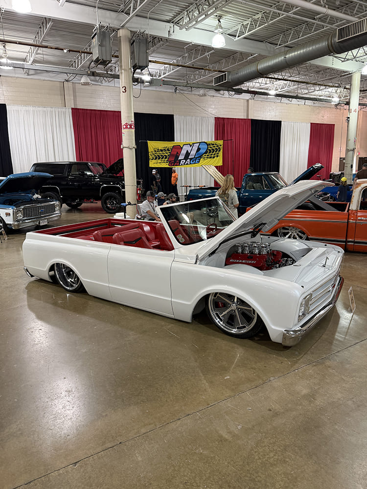 Low-angle front three-quarter view of a custom roofless white Chevrolet C10 pickup featuring red bucket seats, open hood, and eight-stack fuel injection.