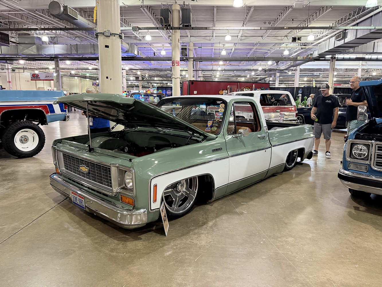 Front three-quarter angle of a slammed light sage green and white two-tone squarebody Chevy pickup with open hood and polished directional wheels on the indoor show floor.
