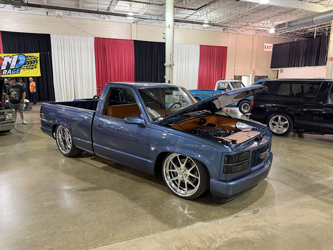 Front three-quarter view of a lowered slate-blue GMC OBS regular-cab pickup with an open hood, black billet grille, and large chrome multi-spoke wheels on display indoors.