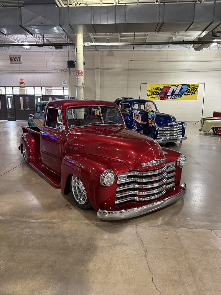 Front three-quarter angle of a slammed metallic burgundy-red vintage Chevy 3100 pickup truck with a chrome grille on display inside an event hall.