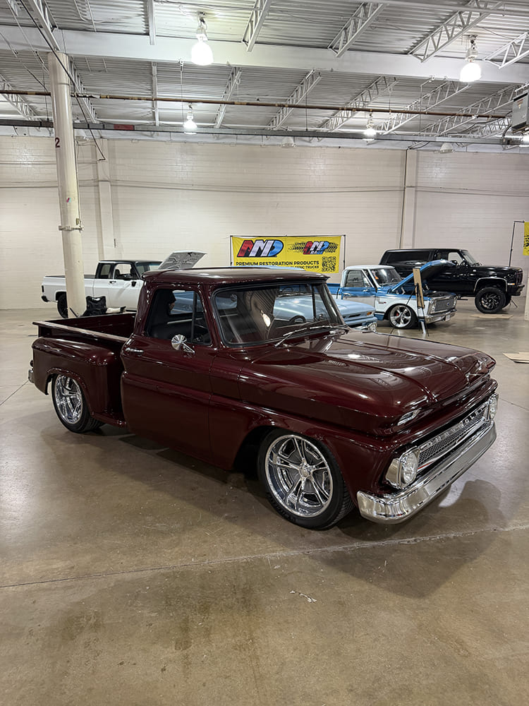 Low-angle front three-quarter view of a dark metallic cherry-red 1960s Chevrolet C10 Stepside pickup truck on chrome multi-spoke wheels indoors.