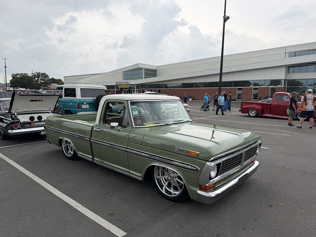 Front three-quarter view of a lowered sage olive and white two-tone Ford F-100 Bumpside pickup truck parked in an outdoor fairgrounds lot.