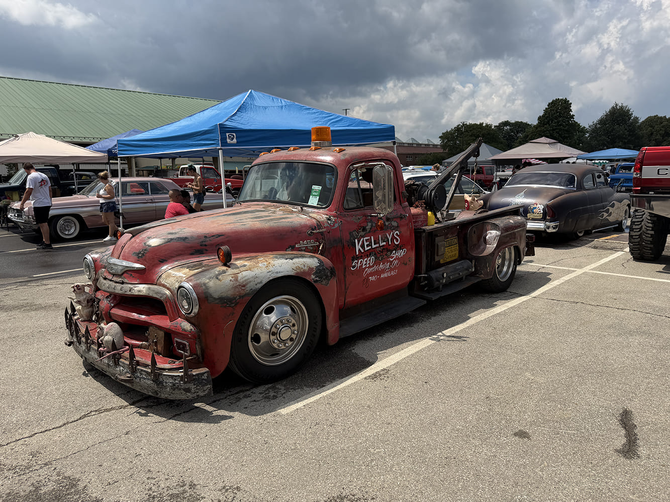 Front three-quarter view of a weathered red Chevy 6400 commercial wrecker truck with boom crane and patina parked under event canopies.