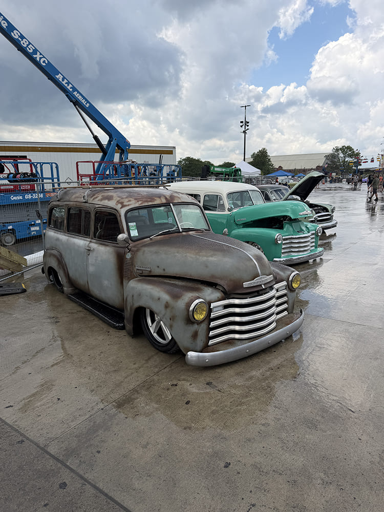 High front three-quarter angle of a rust-patina classic Chevrolet Suburban Carryall wagon parked beside a mint-green vintage truck on a wet tarmac.