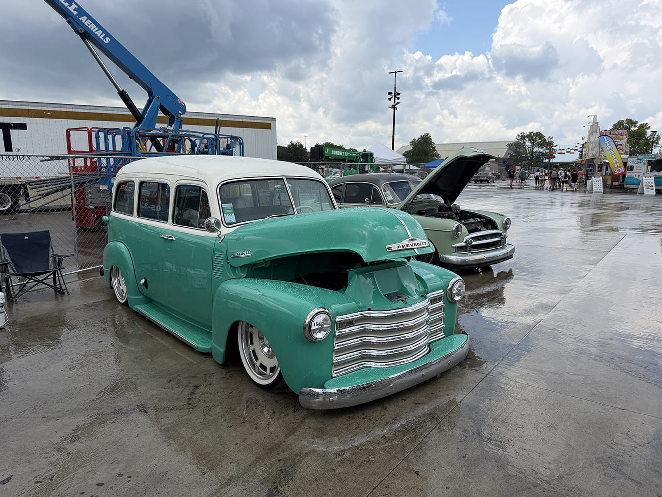 Front three-quarter view of a restored seafoam green and white two-tone Chevrolet Suburban Carryall with an open hood on a wet outdoor concourse.