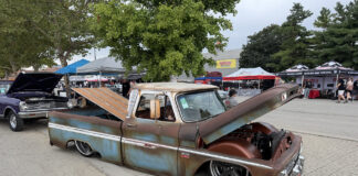 Front three-quarter angle of a slammed weathered Chevy C10 pickup with an open hood, raised wood bed floor, and chrome-rimmed wheels outdoors.