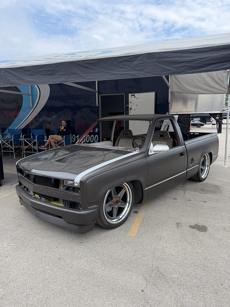 Front three-quarter view of a lowered matte black Chevy C1500 OBS pickup truck build missing windshield glass parked under an event tent canopy.