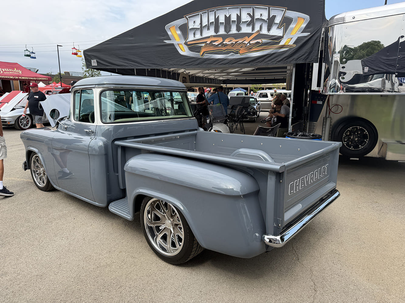 High rear three-quarter view of a custom gloss gray Chevrolet Task Force stepside pickup with polished wheels parked outside an exhibition trailer.