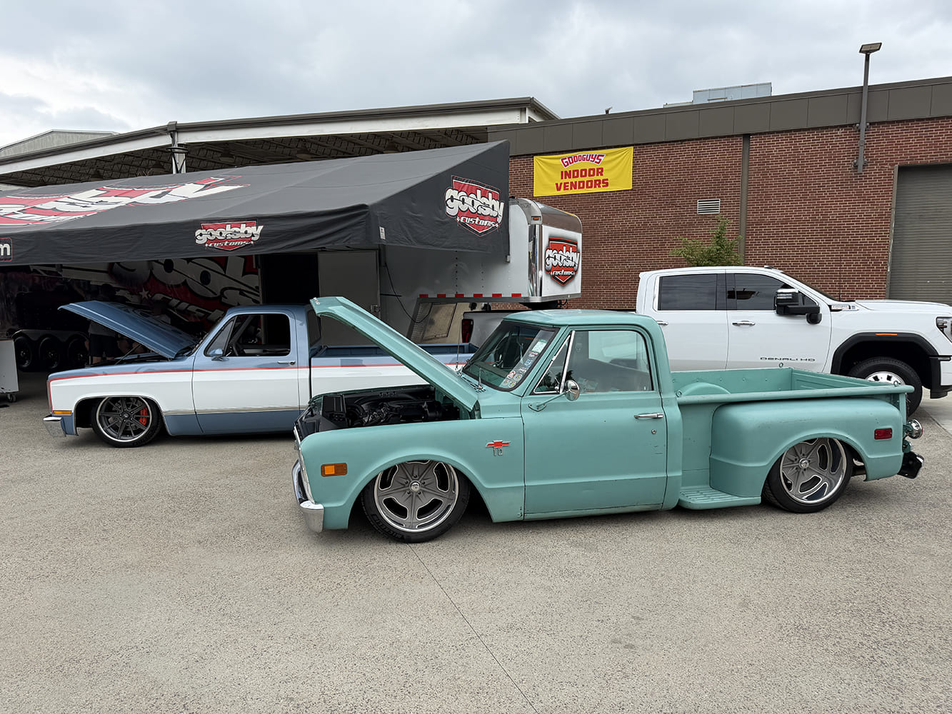 Side three-quarter perspective of a slammed matte green Chevrolet C10 stepside pickup with an open hood parked on asphalt next to a squarebody truck outside a Goolsby Customs event tent.