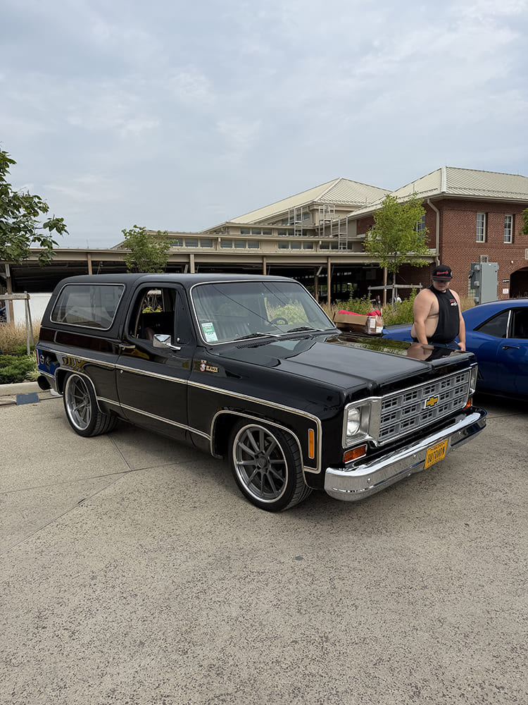 Front three-quarter angle of a clean gloss black vintage Chevrolet K5 Blazer with chrome grille trim and dark gray multi-spoke wheels parked on concrete in front of a brick building.