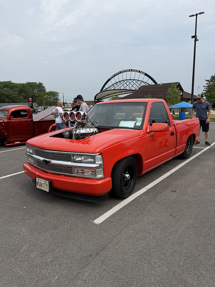 Front three-quarter shot of a bright red Chevrolet C1500 OBS pickup featuring an exposed supercharged V8 engine and black drag wheels parked near the Ohio Expo Center arch.
