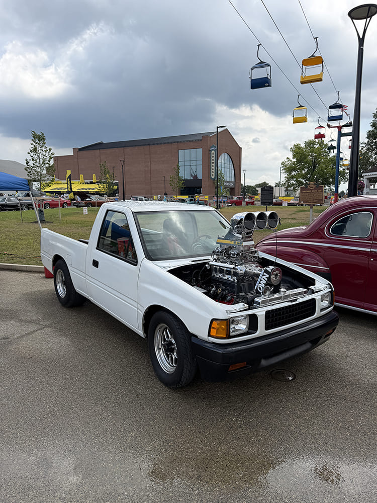 High front three-quarter perspective of a white mini truck featuring a polished supercharged V8 with a tall bug catcher intake scoop towering over the hoodless engine bay beneath skyride cars.