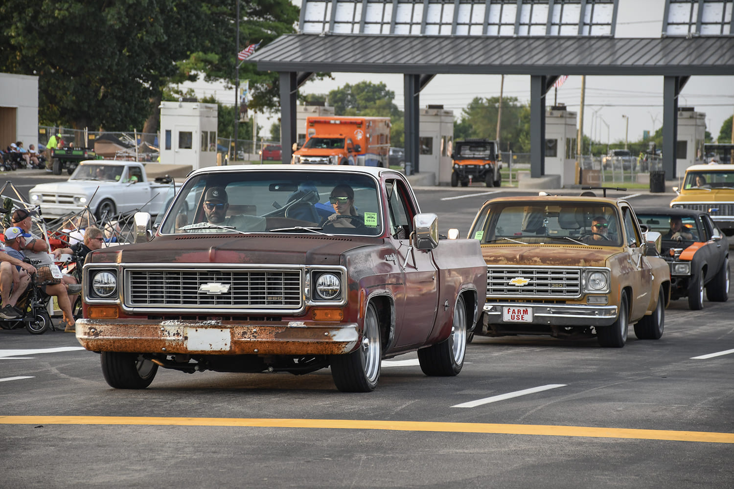 Front three-quarter view of a weathered brown patina squarebody Chevrolet C10 pickup cruising past the entrance gates at the NSRA Street Rod Nationals alongside a line of classic trucks.