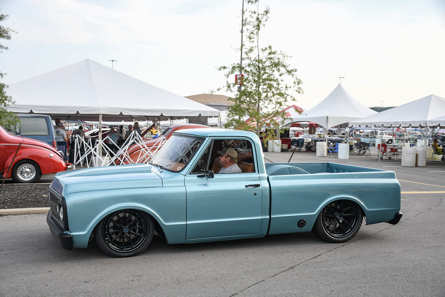 Driver-side profile of a slammed light metallic blue Chevrolet C10 pickup rolling past outdoor event tents on black modern multi-spoke wheels.